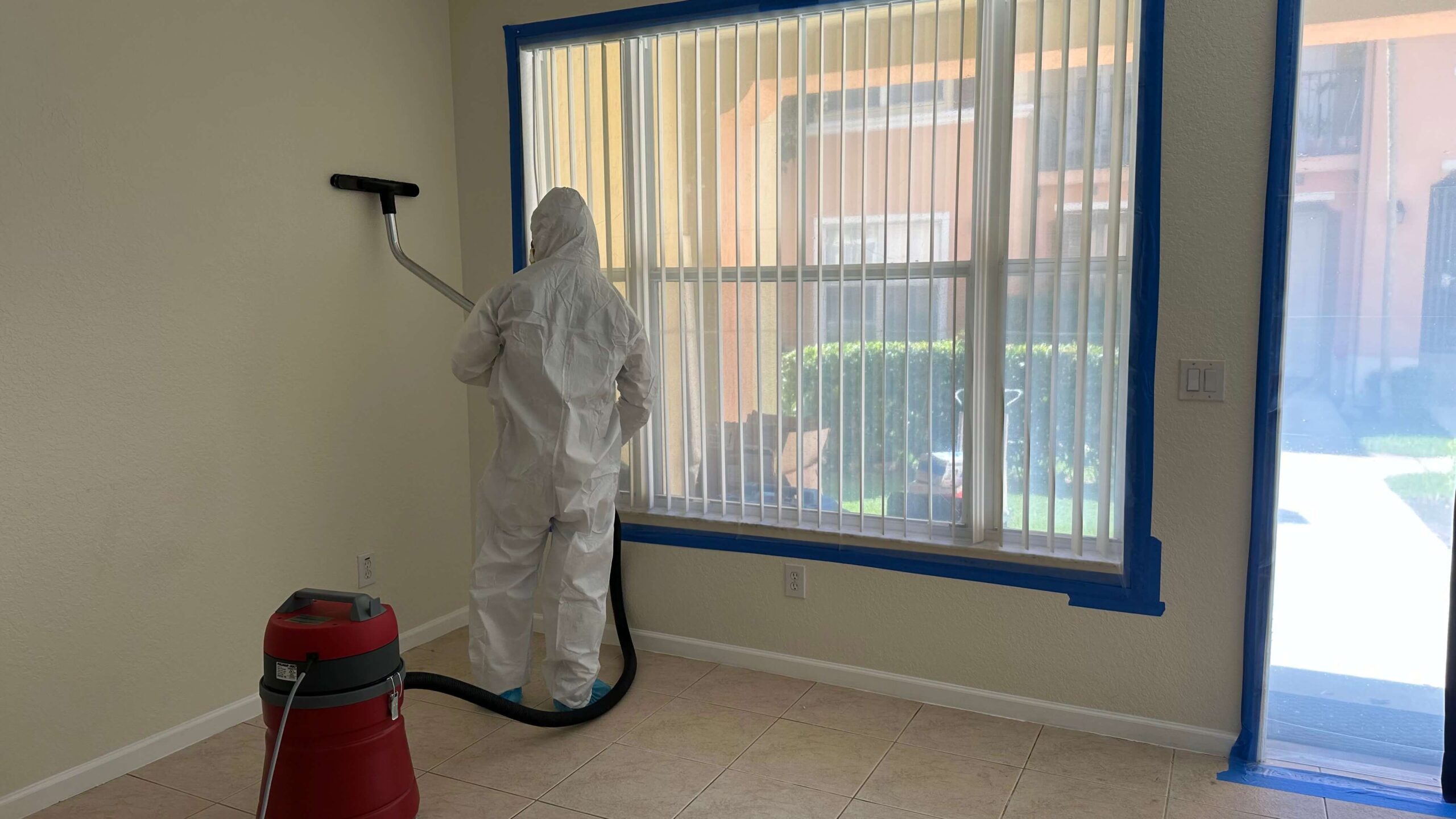 worker-cleaning-mold-on-wall A person in a white suit is cleaning a room, focusing on removing mold from surfaces.