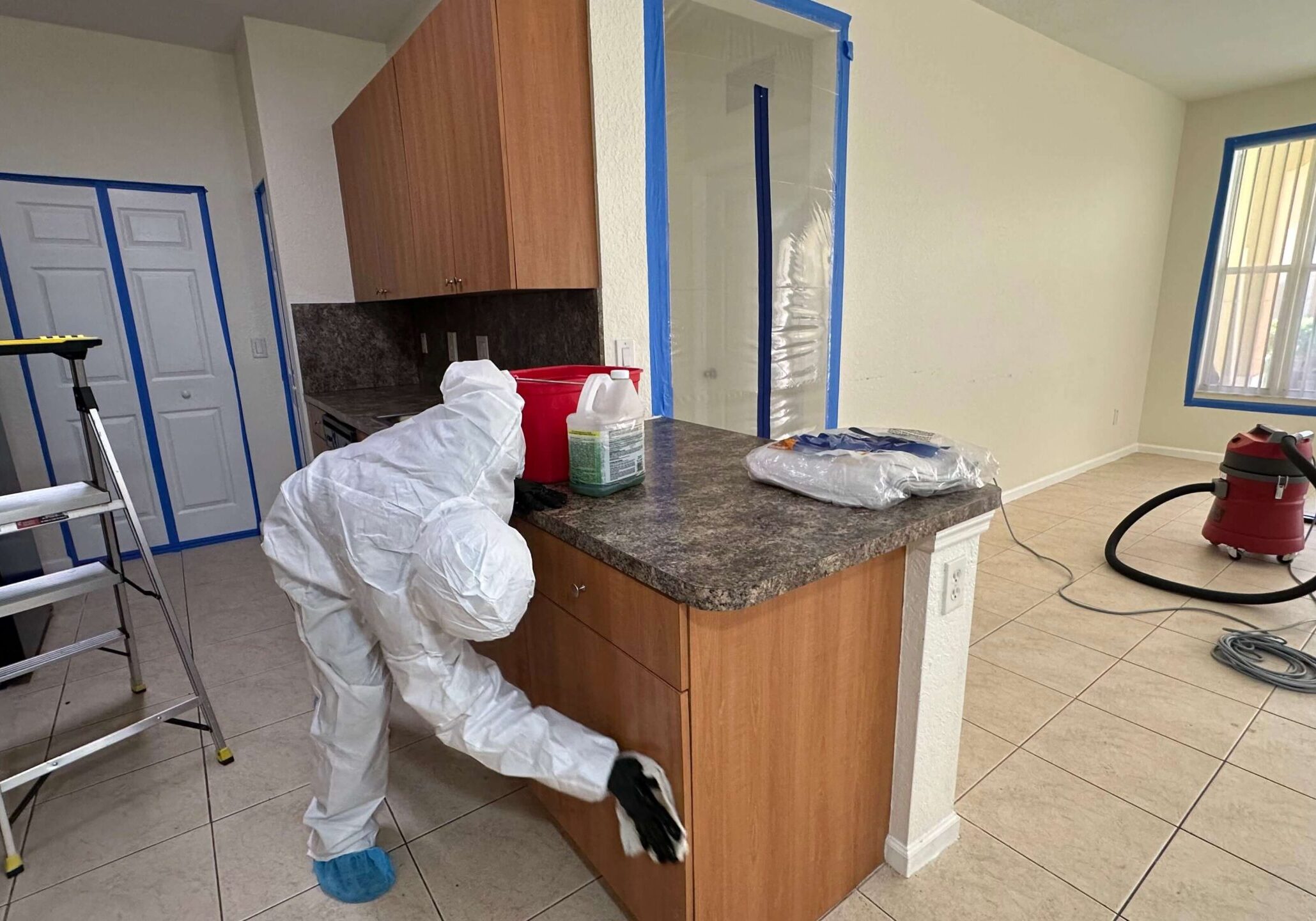 man-removing-mold-from-kitchen-counter A person in a white suit scrubs mold from surfaces in a kitchen, ensuring a clean and safe environment.