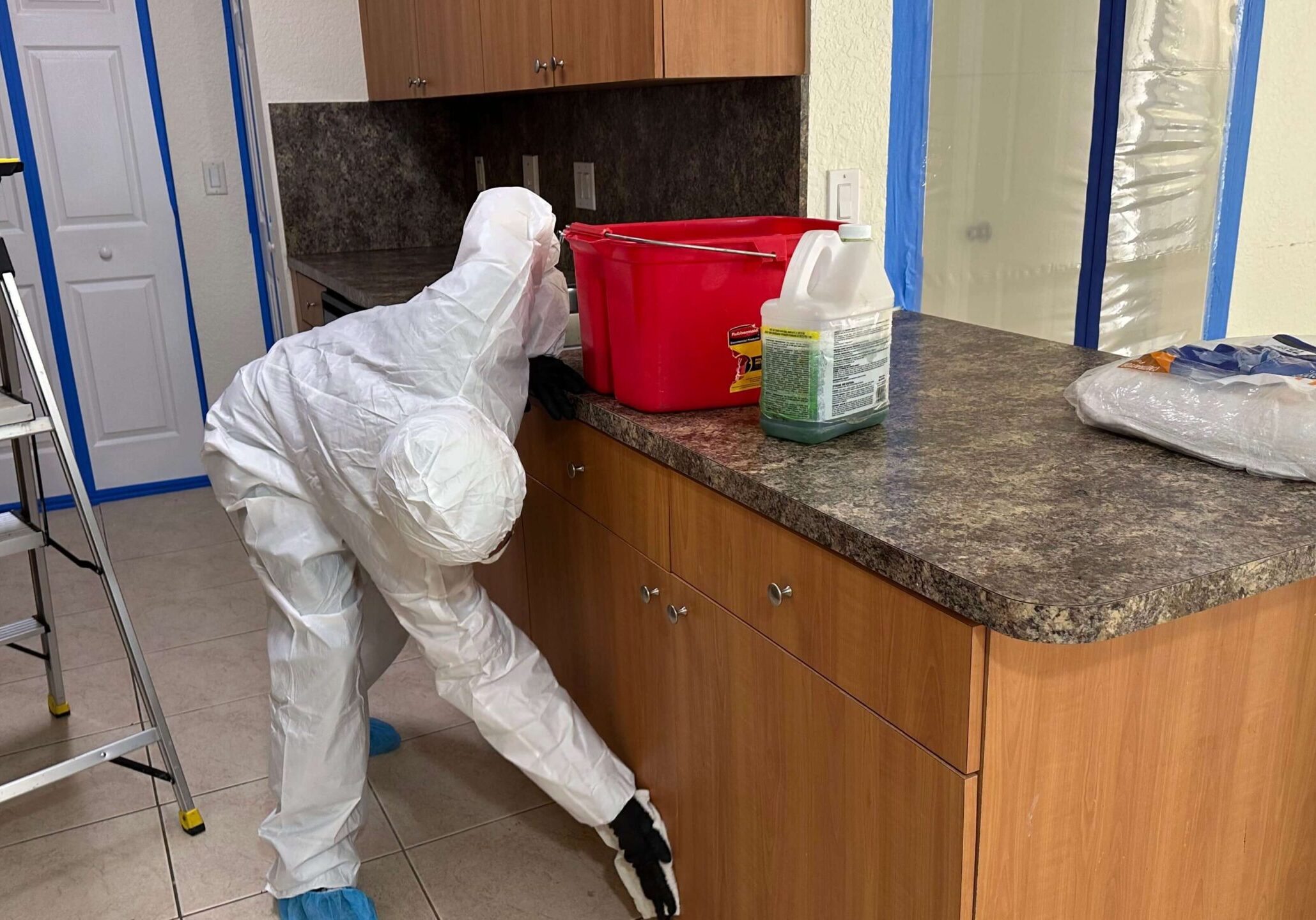 worker-cleaning-kitchen-counter A person in a white suit scrubs a kitchen counter, removing mold and ensuring a clean, safe environment.