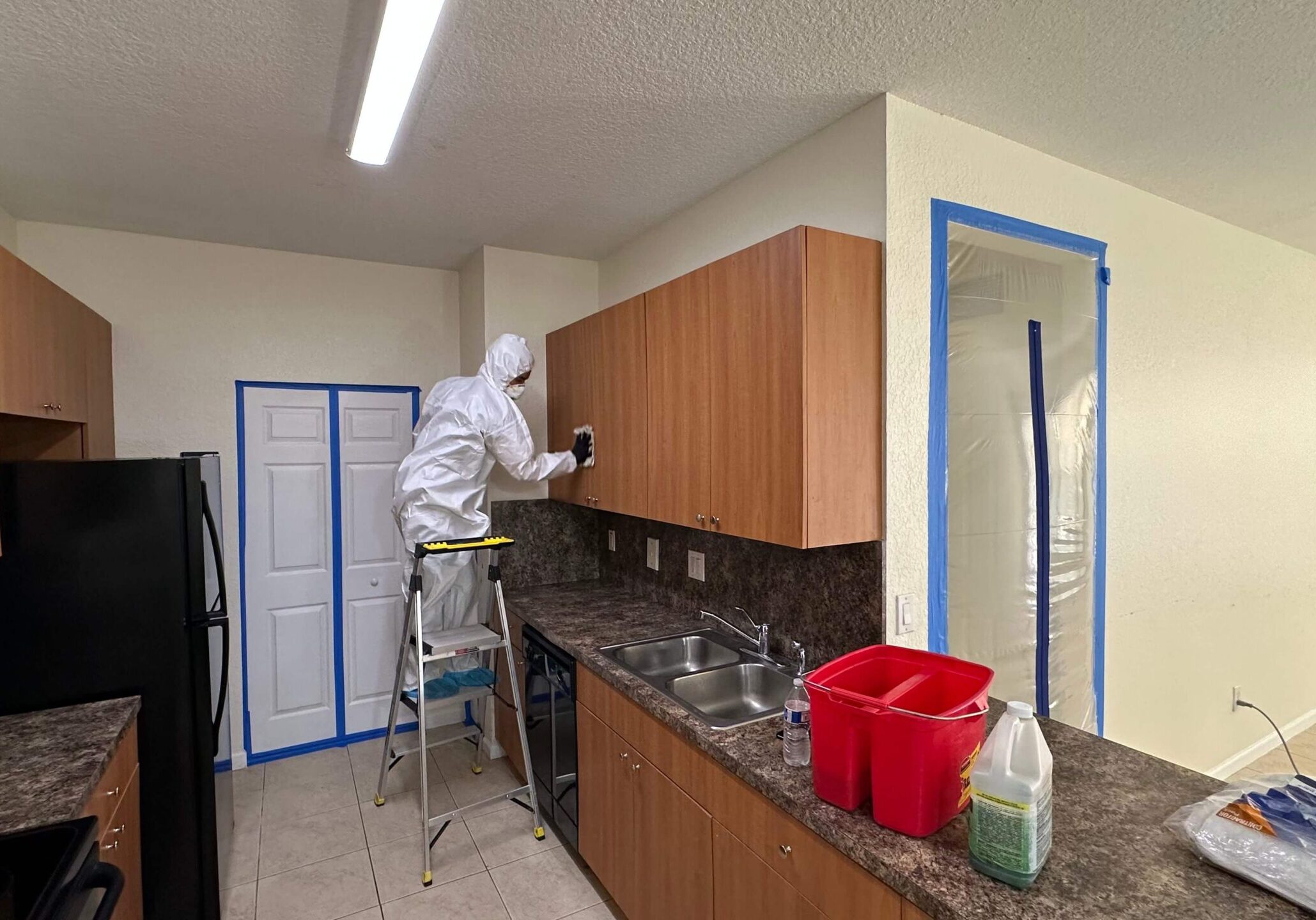 worker-cleaning-mold-on-kitchen-cabinet A man in a white suit is painting a kitchen while addressing mold cleaning.